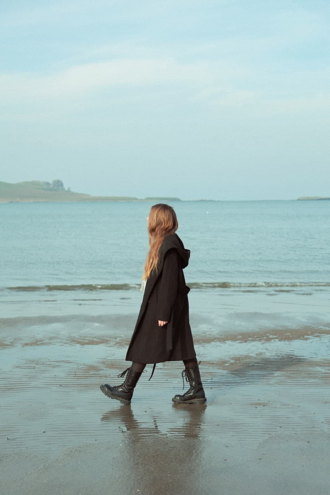 Walking the wet sand at low tide in a long black coat, a headland on the horizon