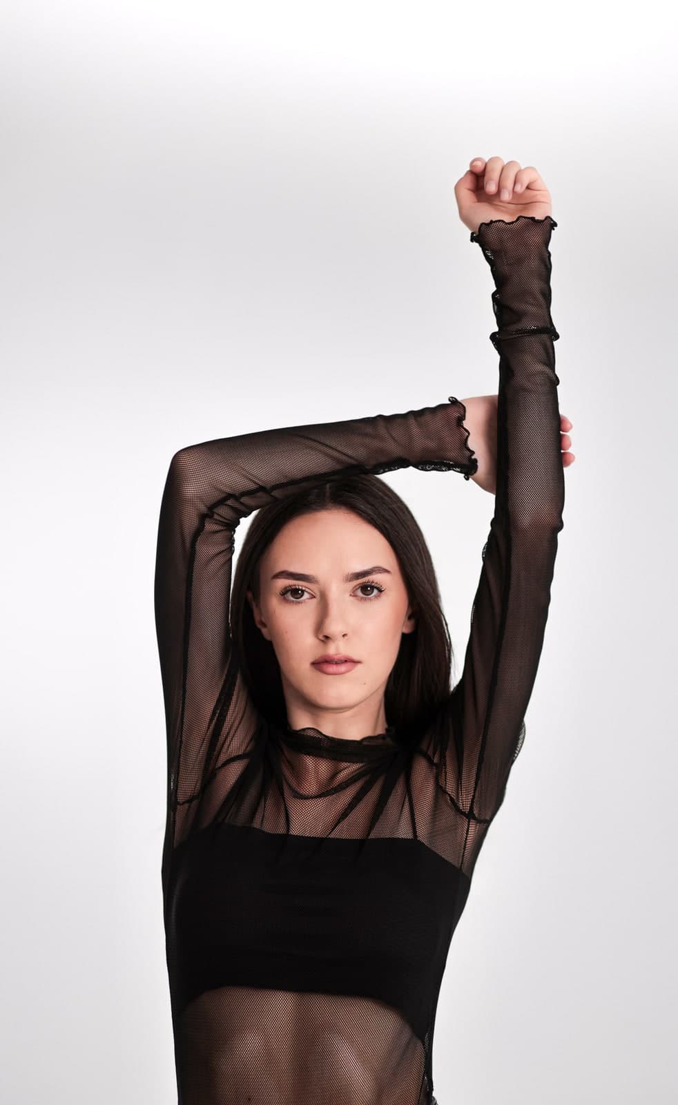 Studio portrait with both arms raised overhead, in a sheer black top on a white backdrop