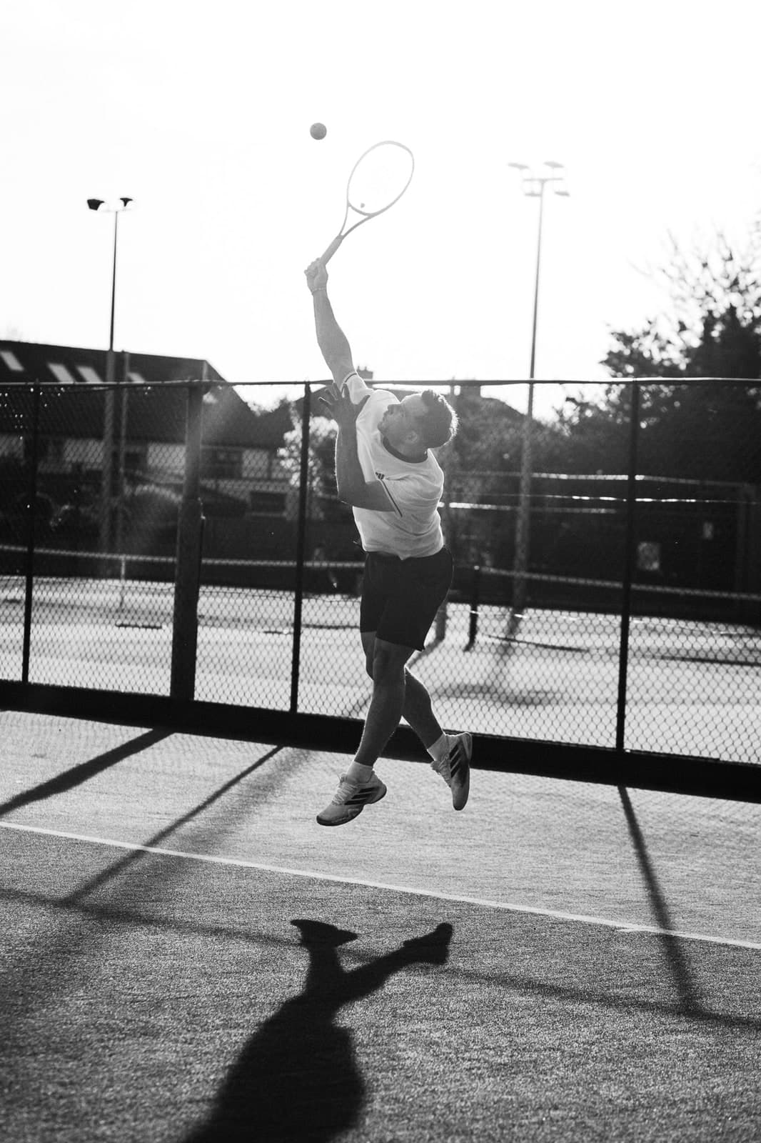 Black-and-white: leaping into a serve on a city tennis court, a long shadow below