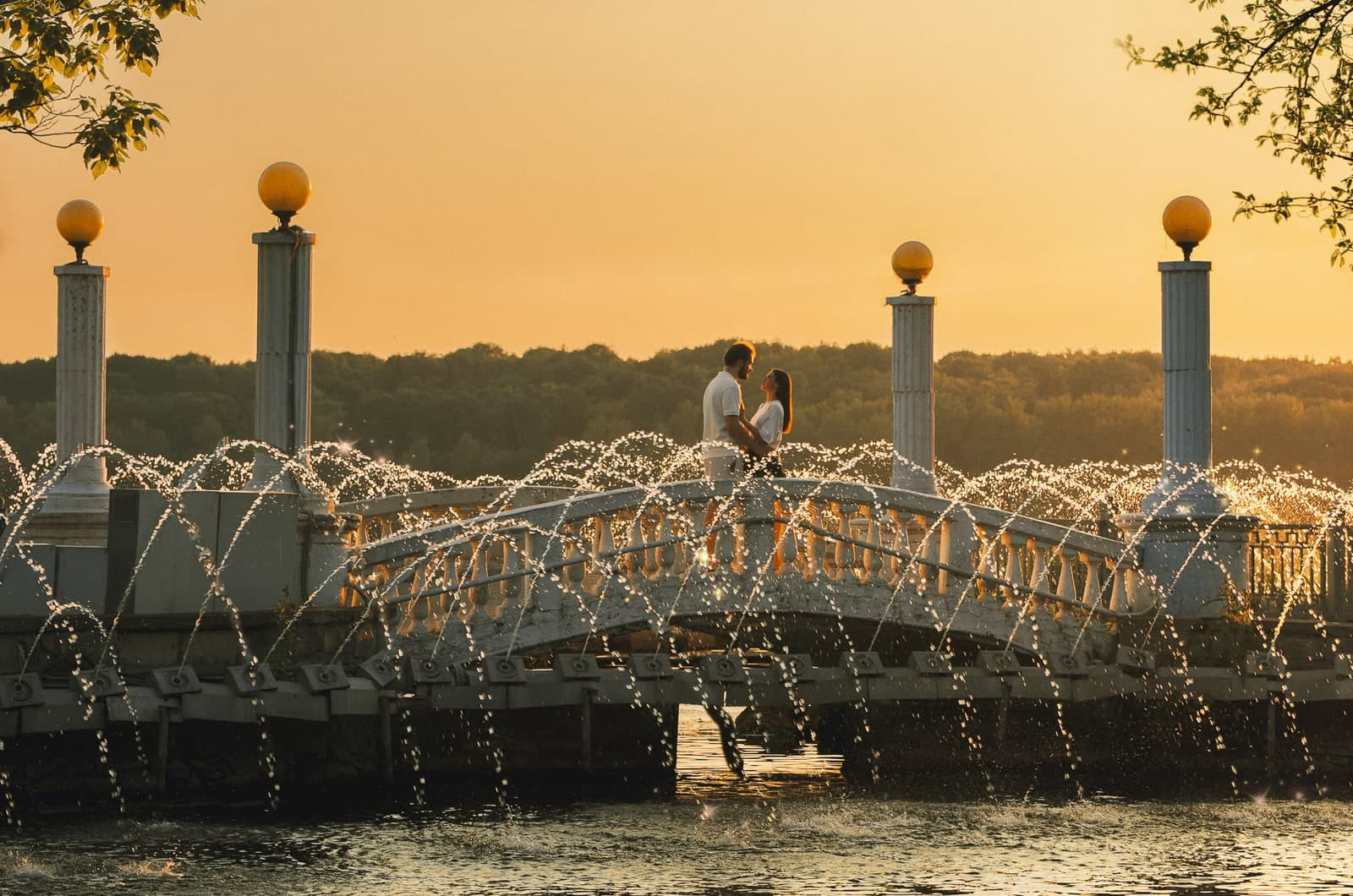 A couple on a bridge behind fountain jets at sunset