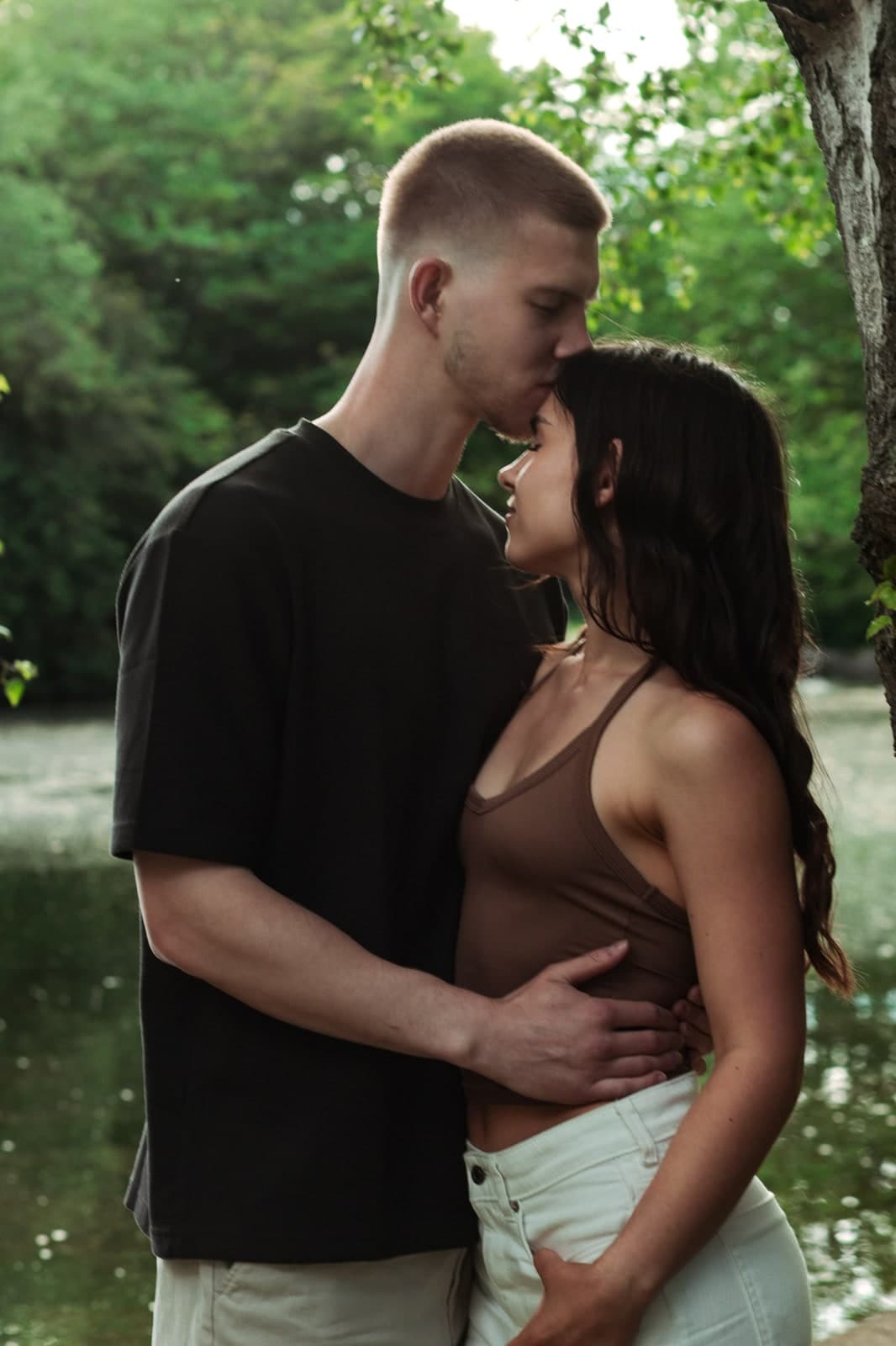 A kiss on the forehead by a pond, in the shade of the trees