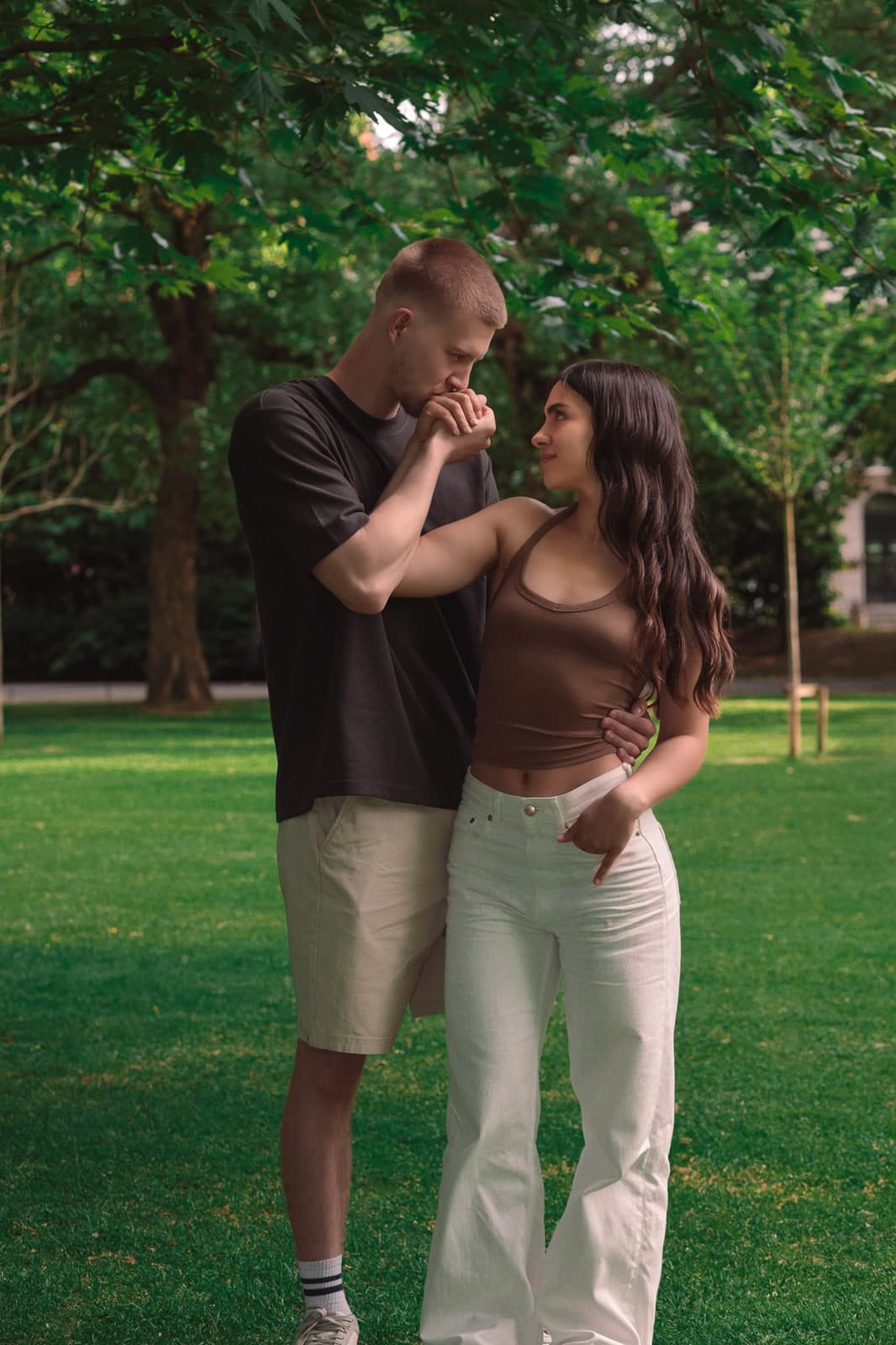 A kiss on the hand on a green lawn under tall trees, both smiling