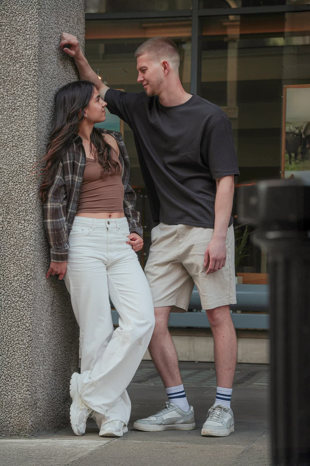 Couple leaning against a shopfront pillar on a city street, trading glances