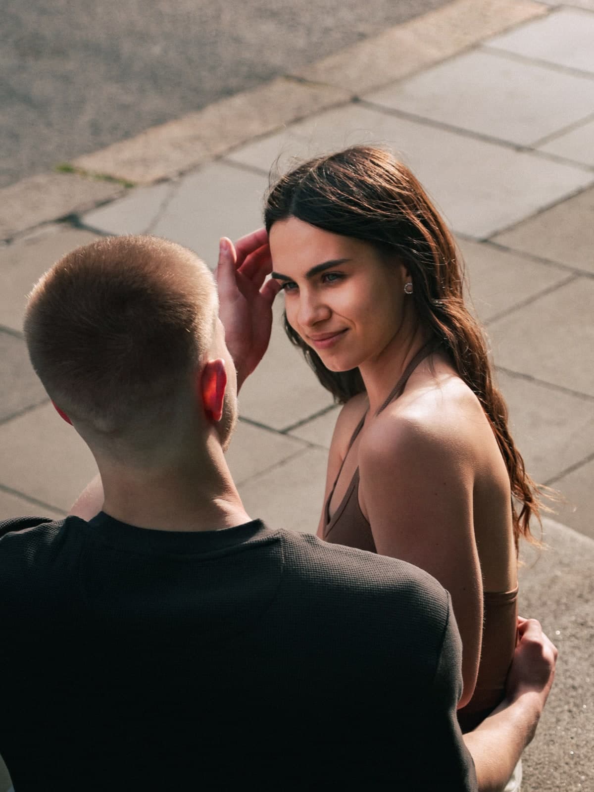Sitting close on a sunny pavement, one tucking hair behind an ear and smiling