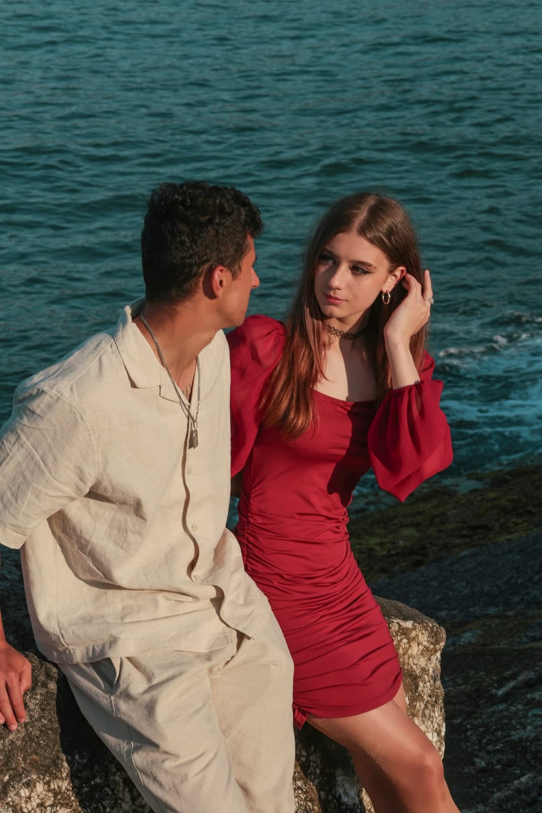 Couple sitting on seaside rocks, in a red dress and a linen shirt
