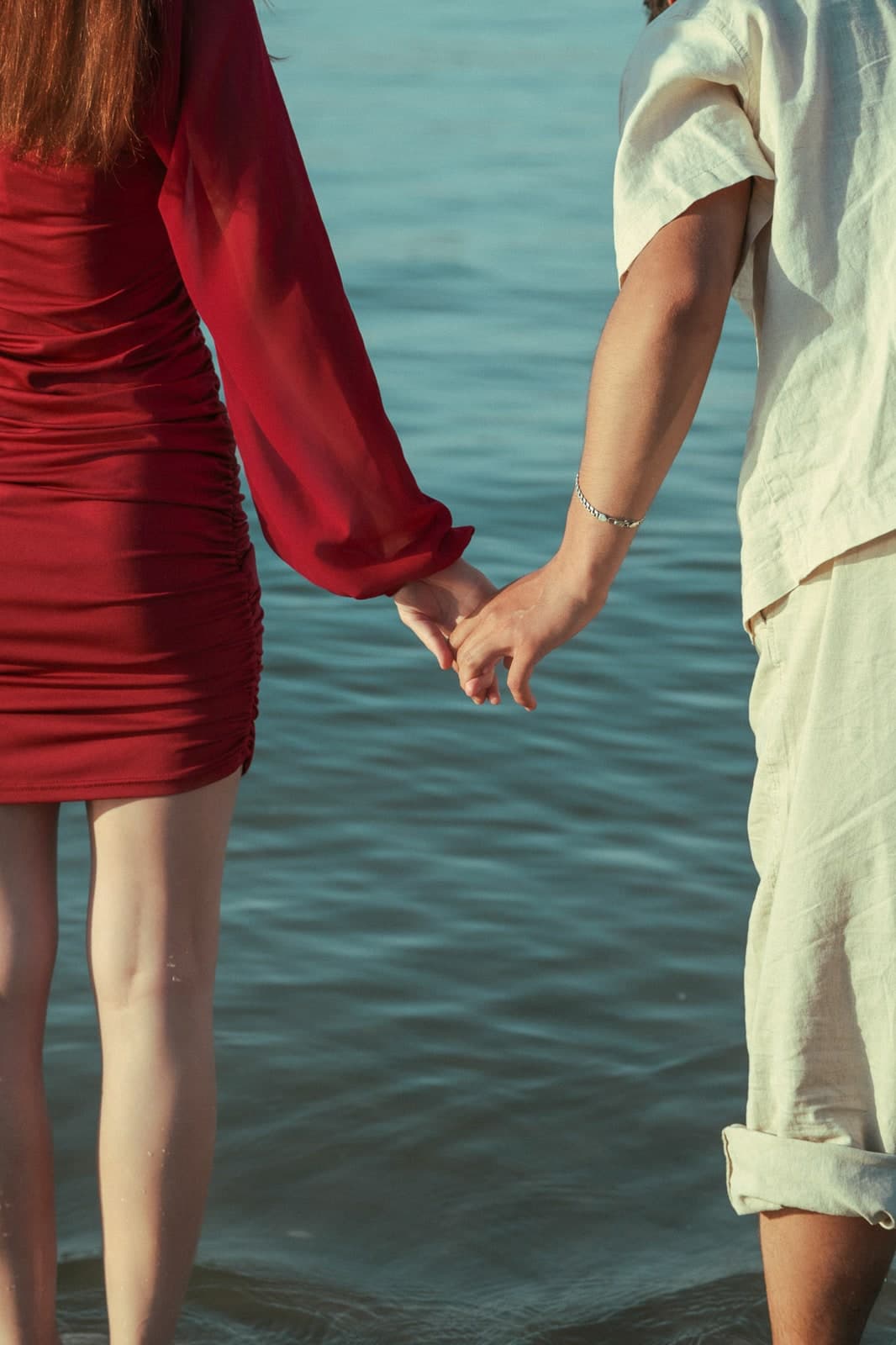 Hands joined at the water's edge, a red dress and rolled-up linen trousers