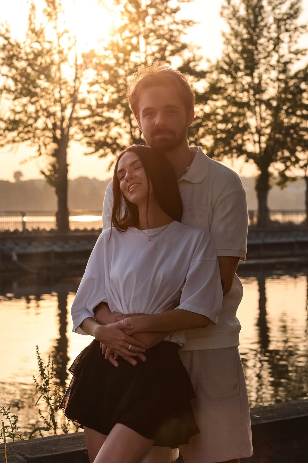 A hug from behind by a lake at sunset, trees in silhouette