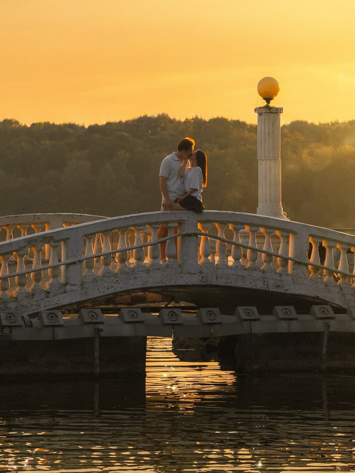 Couple kissing on a white balustraded bridge over a lake at sunset