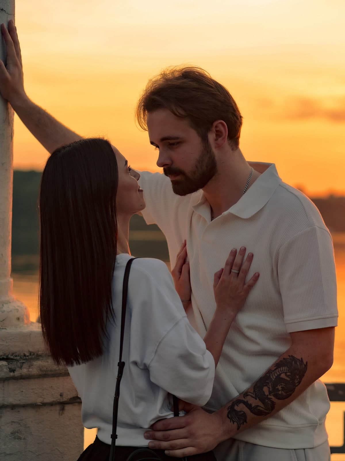Couple close together by a stone pillar at sunset, warm light on the water