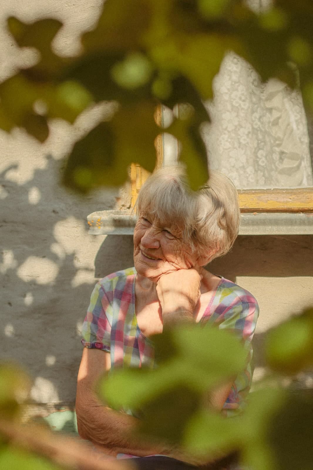 Smiling through vine leaves by a sunlit window, chin on hand