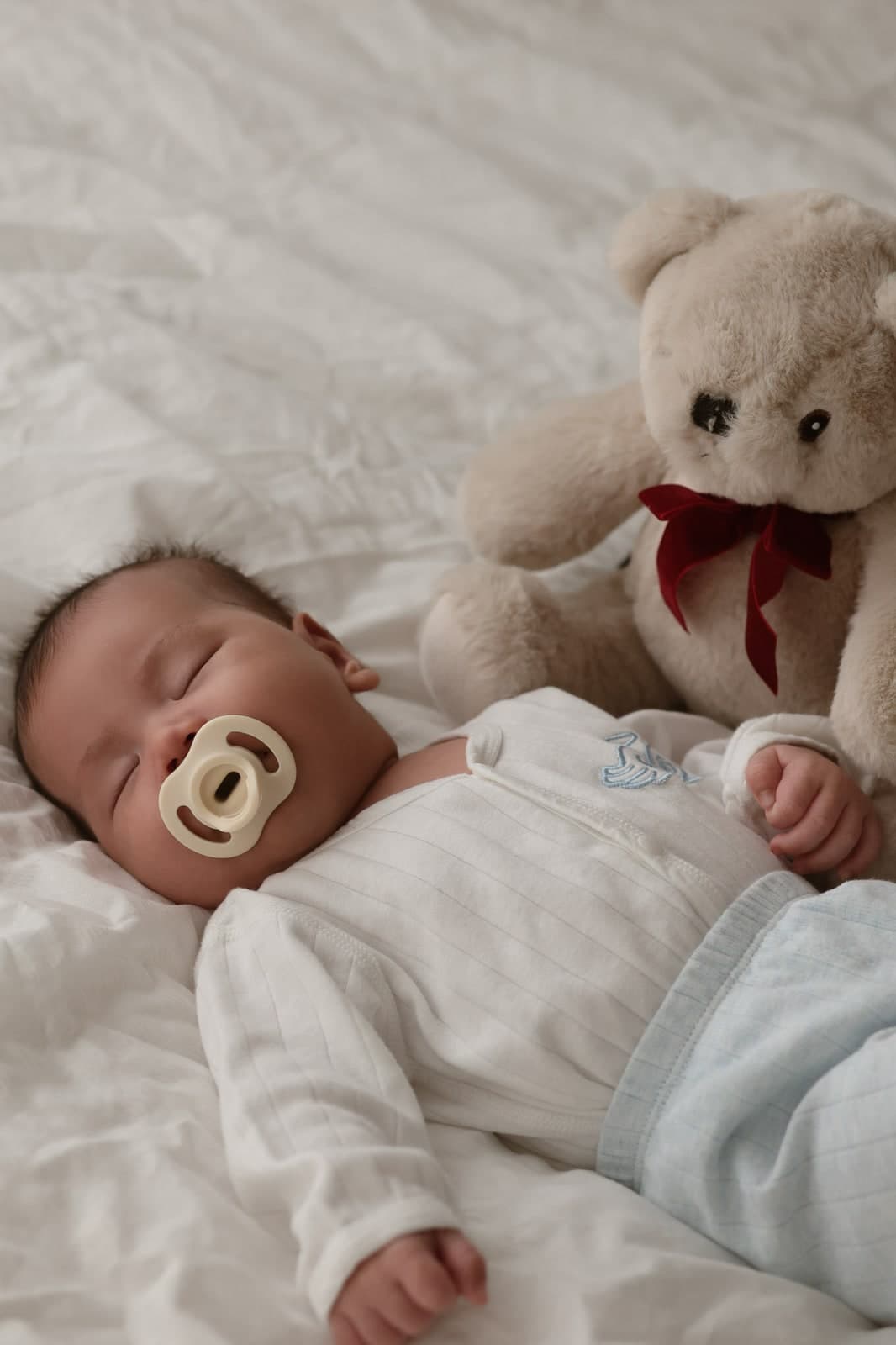 A newborn asleep on white sheets with a dummy, a teddy bear with a red bow alongside