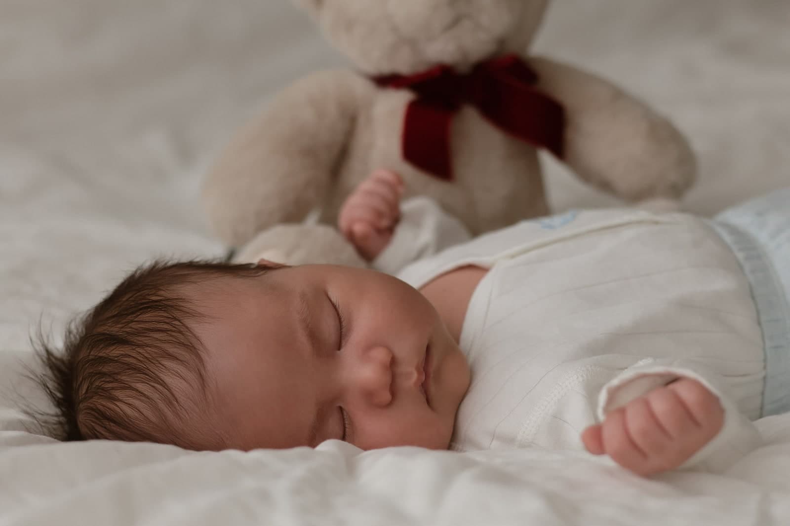 A newborn asleep on a white bed beside a teddy bear