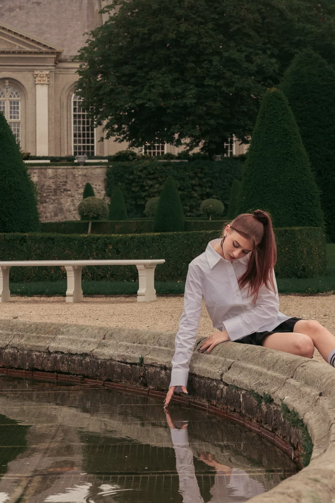 Fingertips in a stone fountain pool, a stately house and clipped yews behind