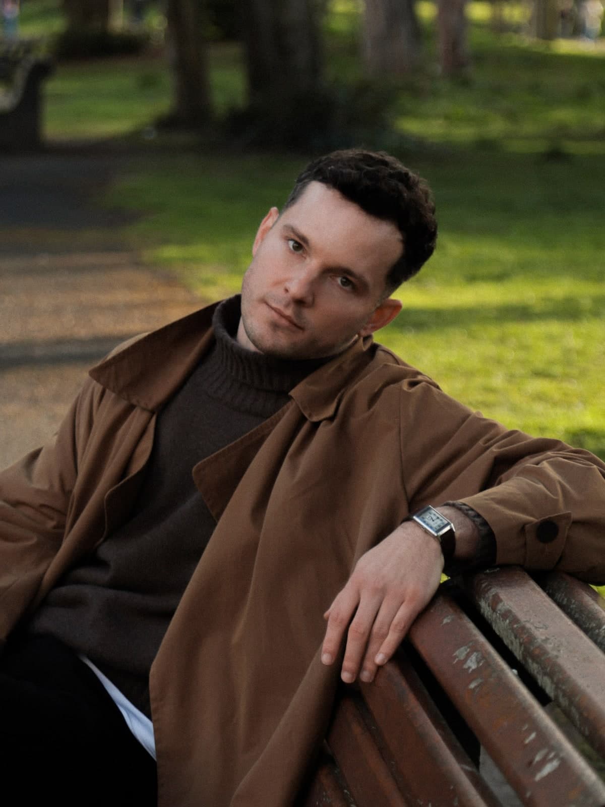 Leaning back on a wooden park bench in a brown coat and roll-neck, late sun on the grass