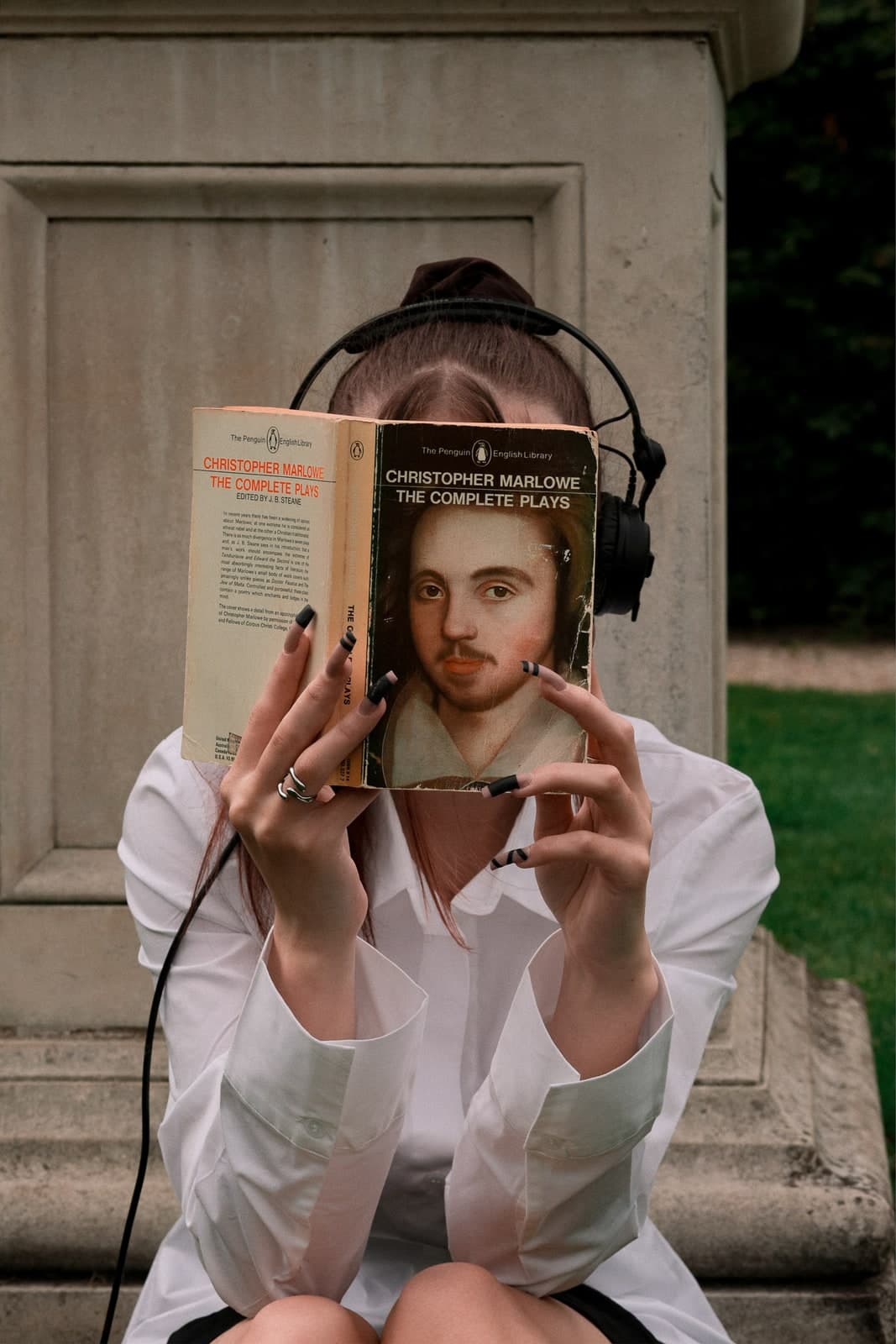 Face hidden behind a Christopher Marlowe paperback, headphones on, by a stone plinth