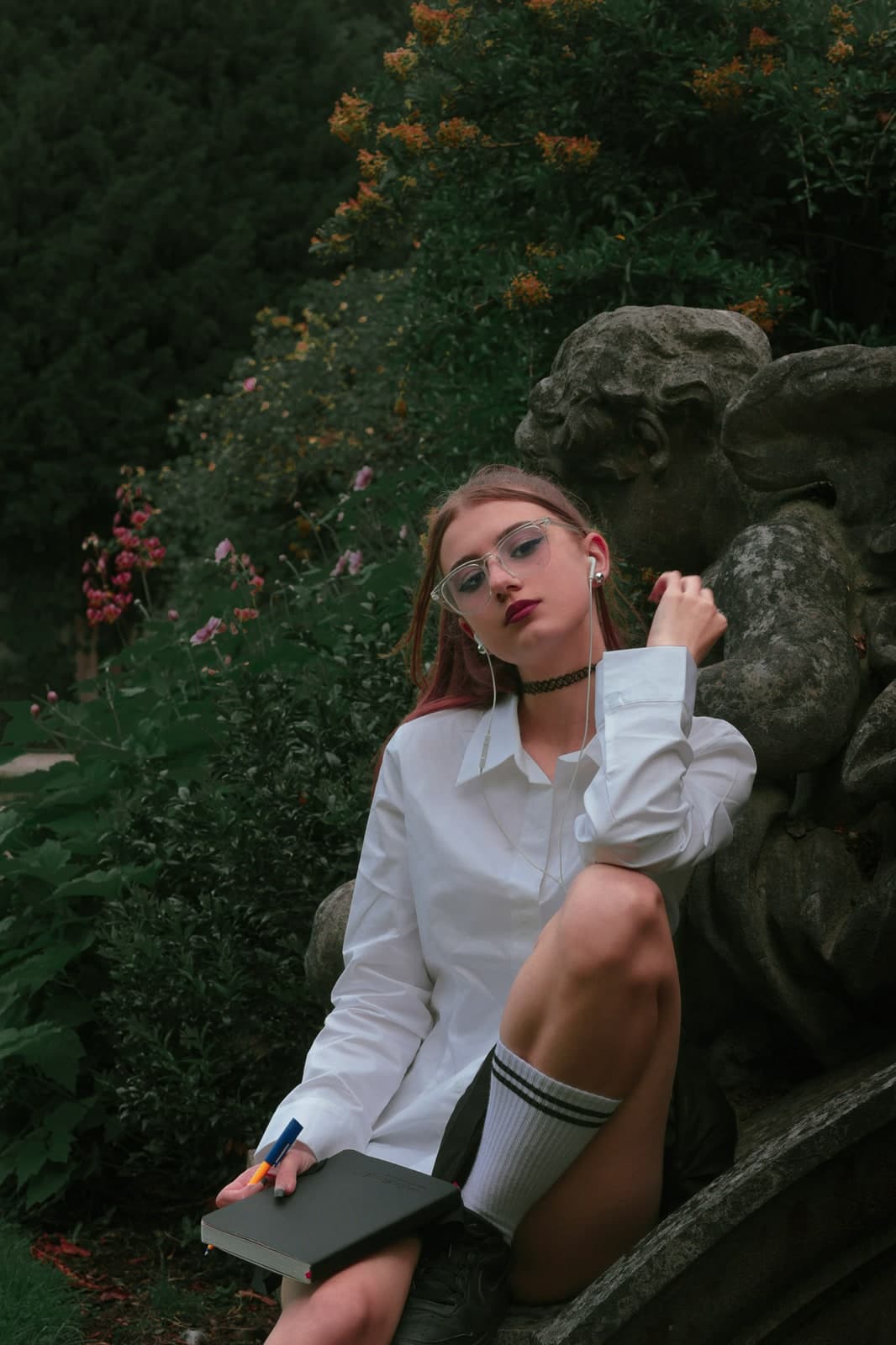 Leaning on a weathered stone statue in round glasses, a notebook in hand, flowers behind