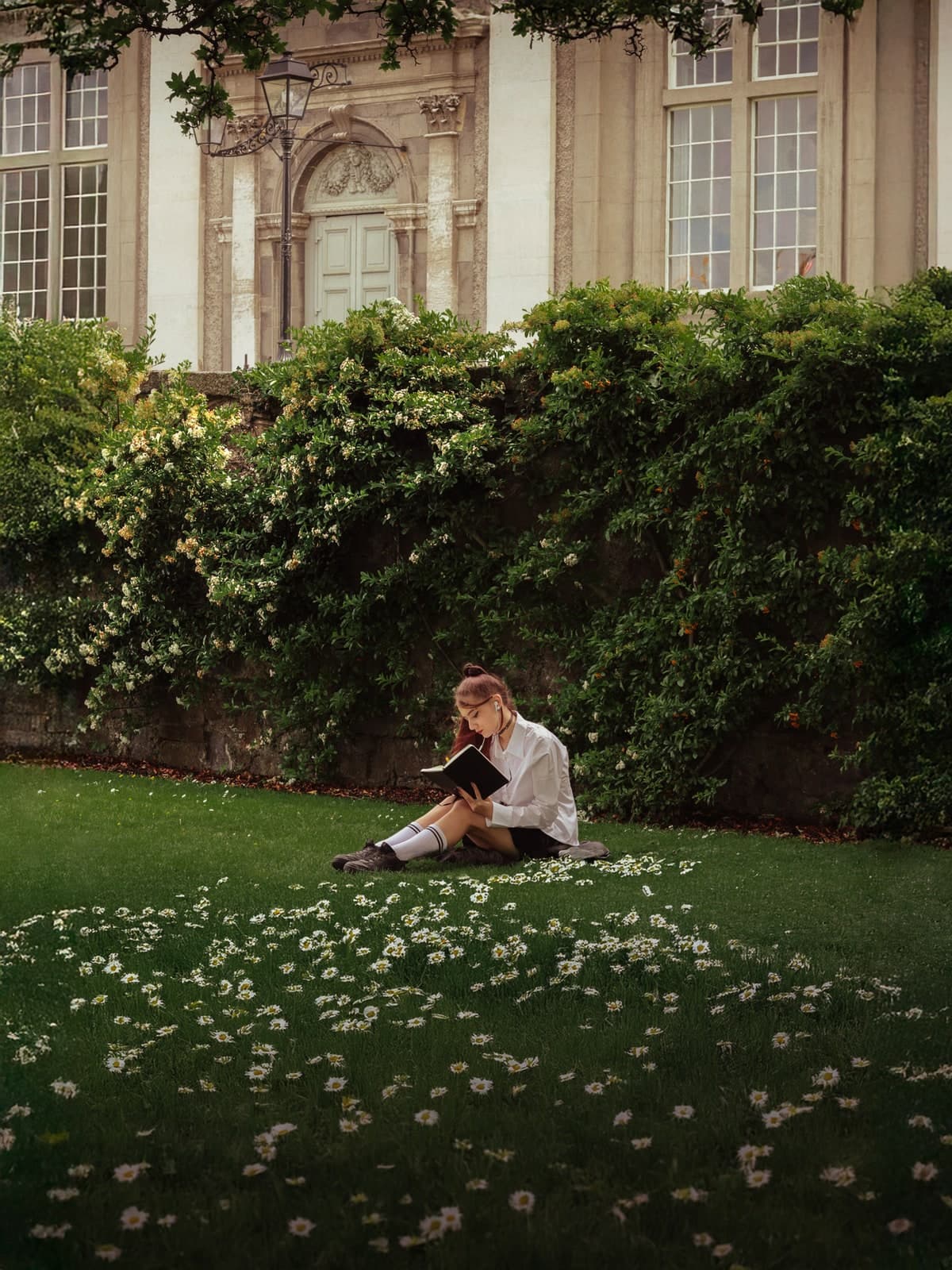 Reading on a lawn of daisies below an ivy-covered wall and a stately house