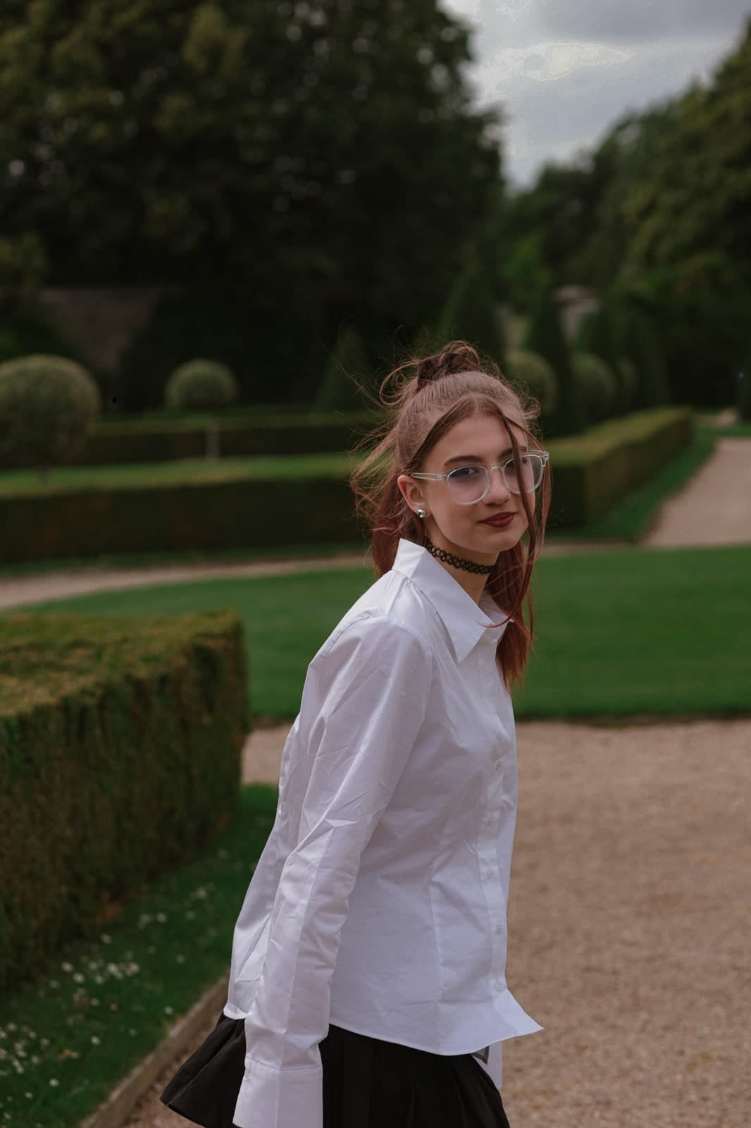 Glancing back on a gravel path between clipped hedges, in a white shirt and glasses