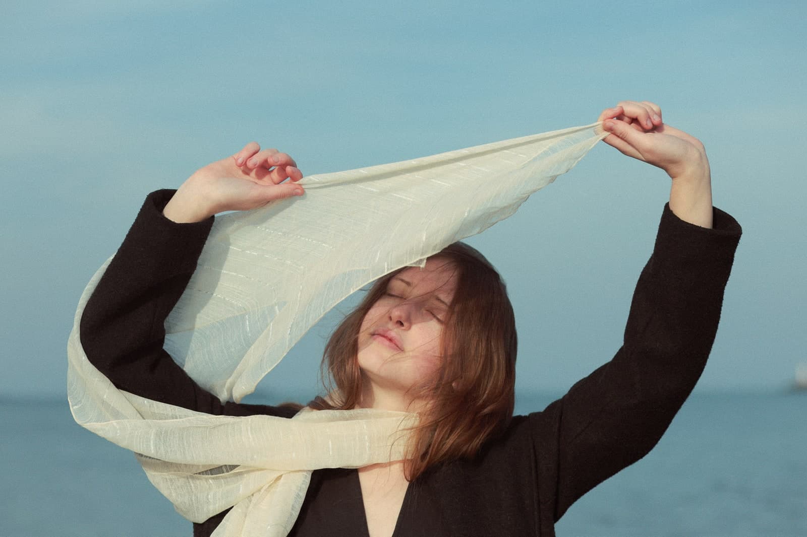 Holding a pale scarf up to the wind with eyes closed, the sea behind