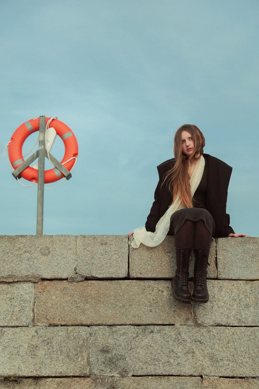 Sitting on a granite sea wall beside an orange lifebuoy, in a black coat and cream scarf
