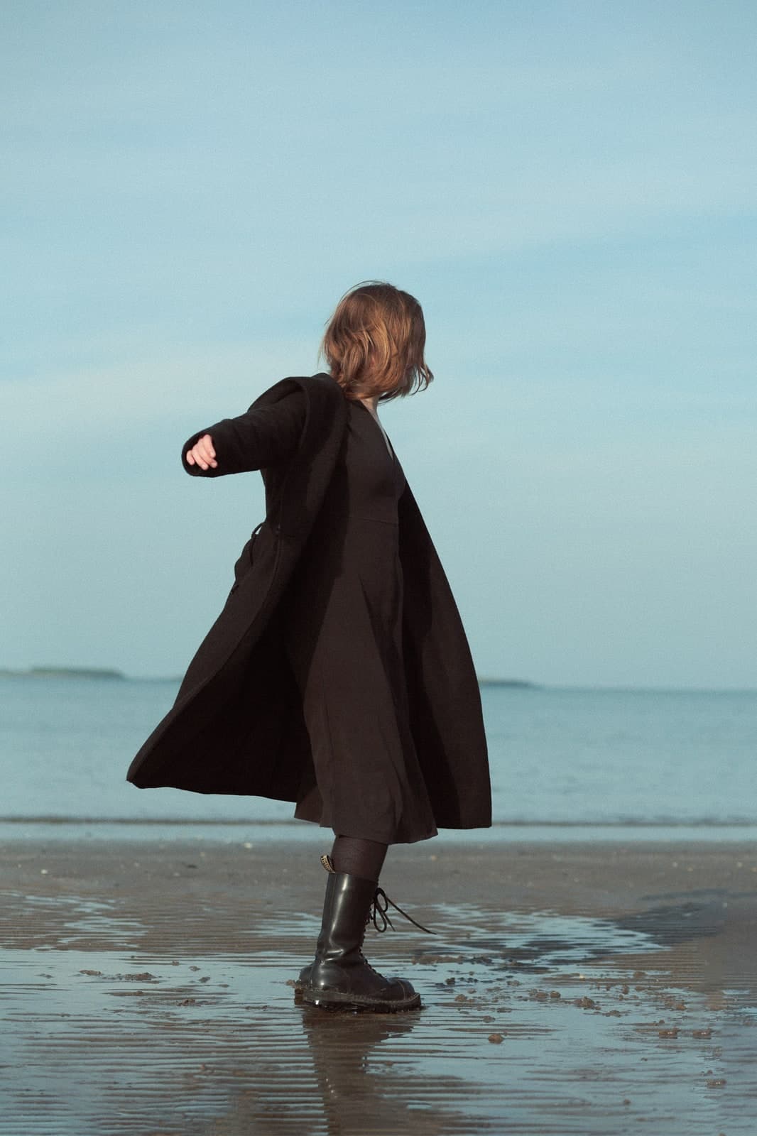 Turning on the wet sand in a long black coat, the sea and a pale sky behind