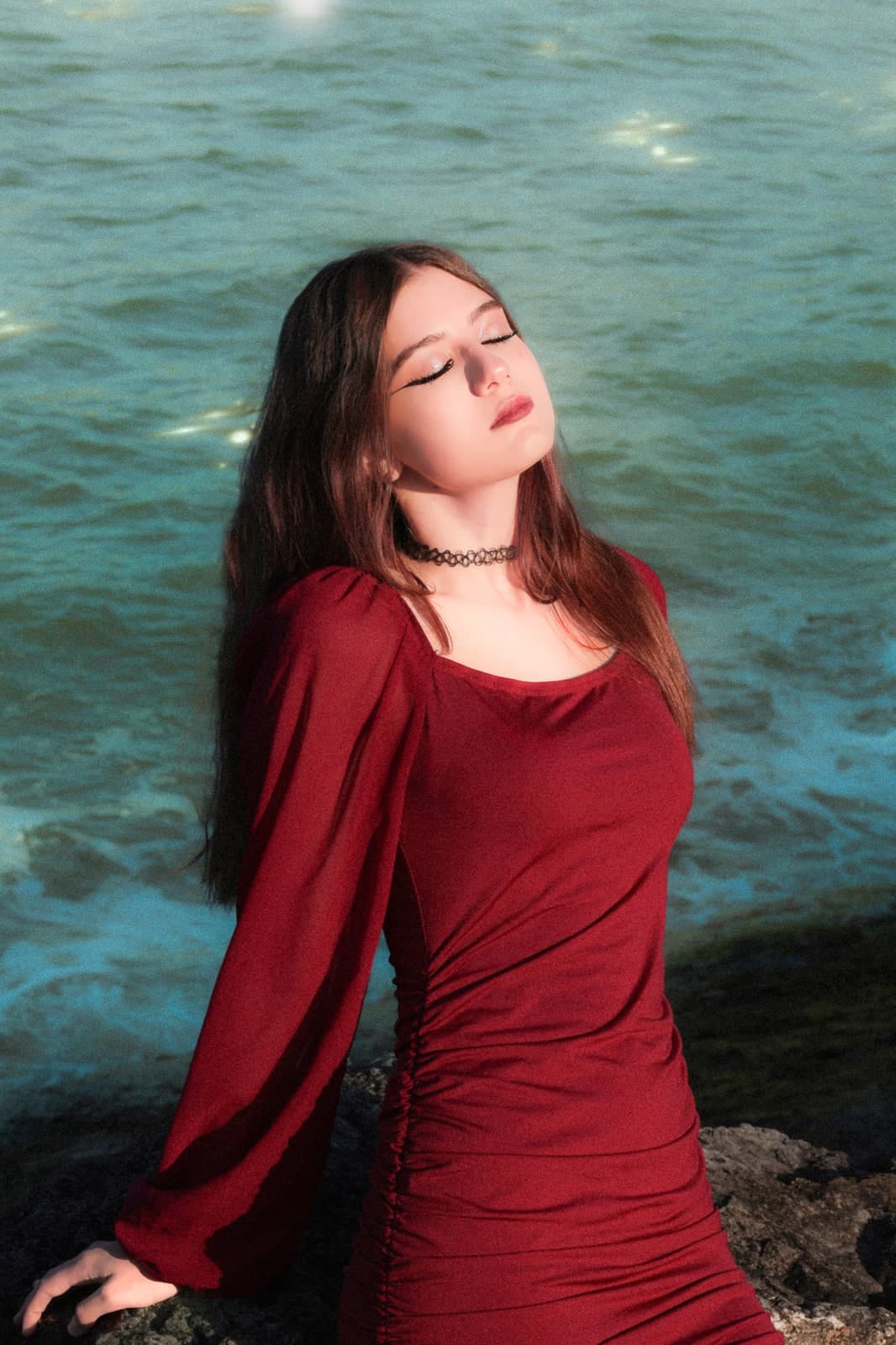 Eyes closed in the sun in a red dress, waves breaking on the rocks behind