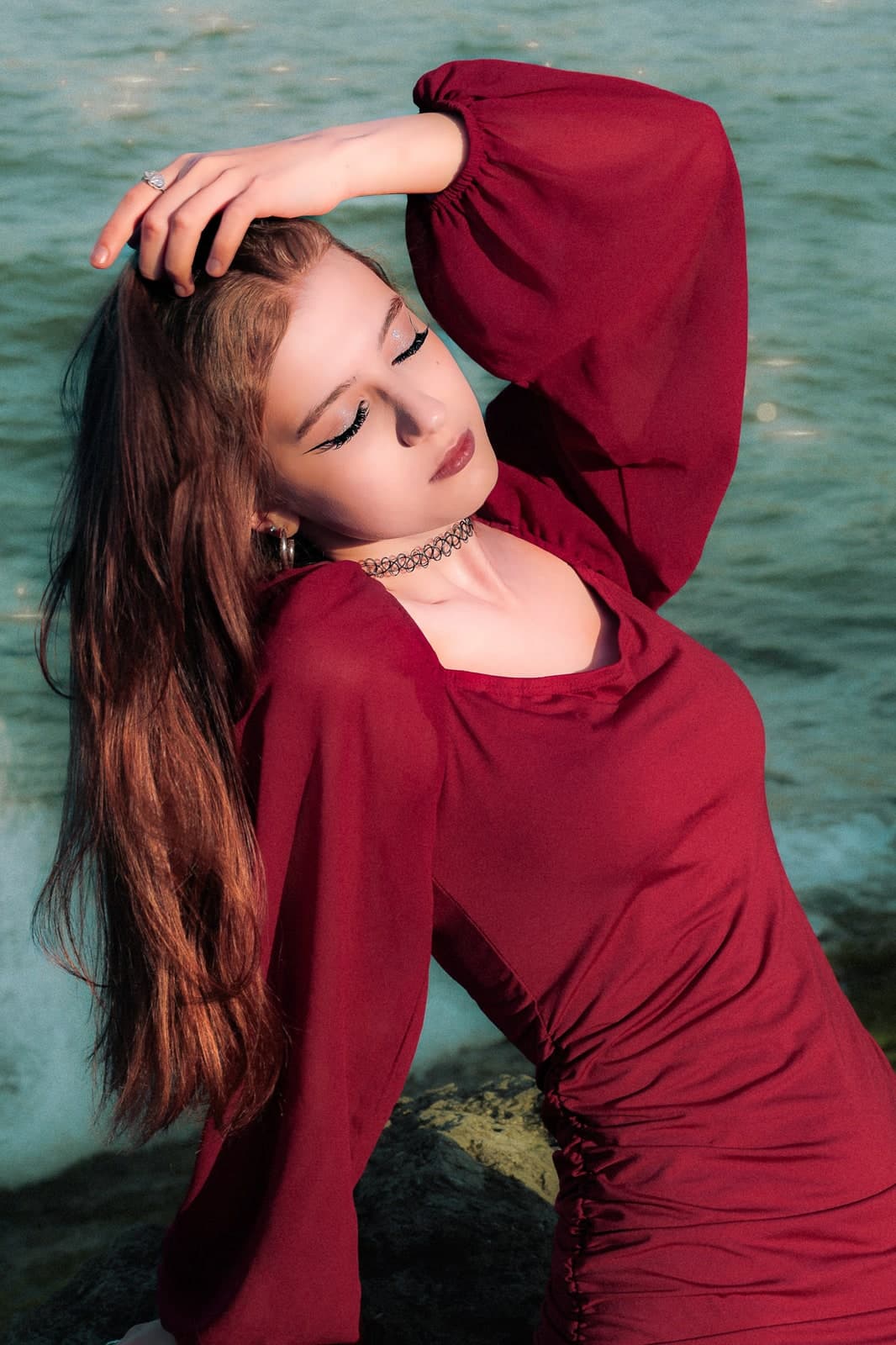 A hand in long hair, eyes closed, in a red dress by the sparkling sea