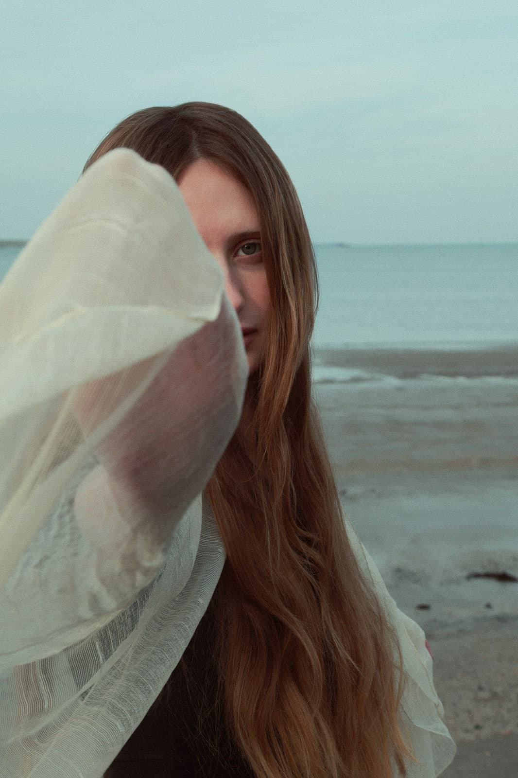 Long hair and a sheer white sleeve drawn across the face on an empty beach
