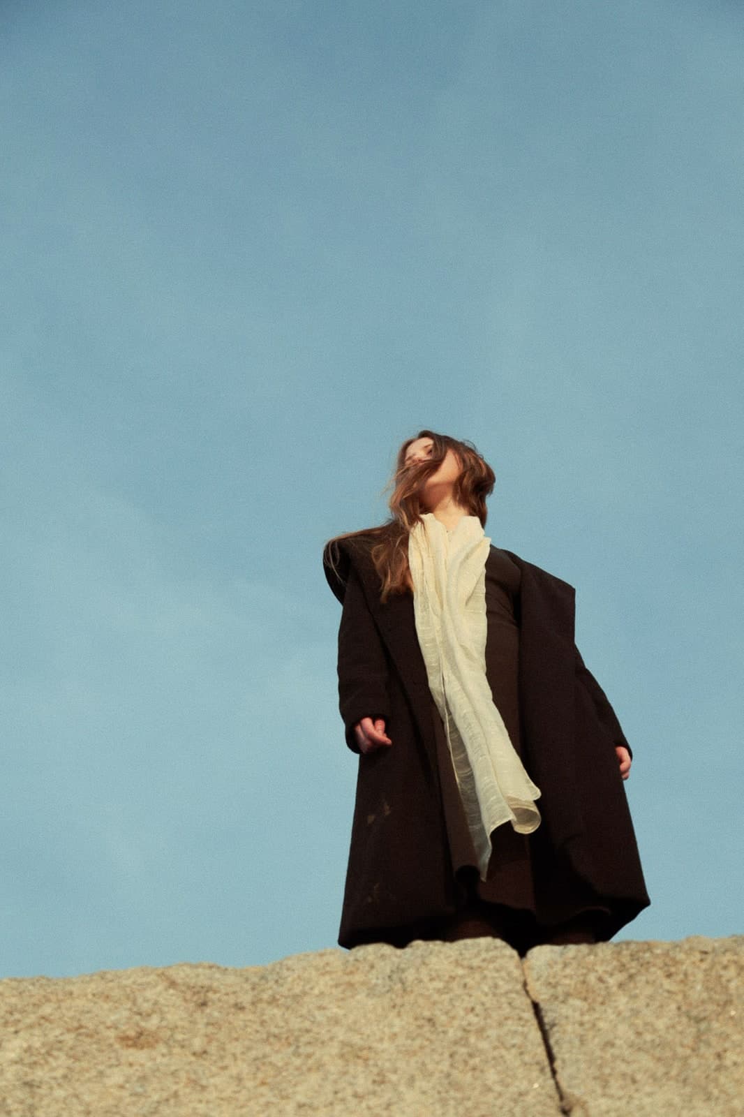 Standing on a granite rock against the sky, head tipped back, in a black coat and cream scarf