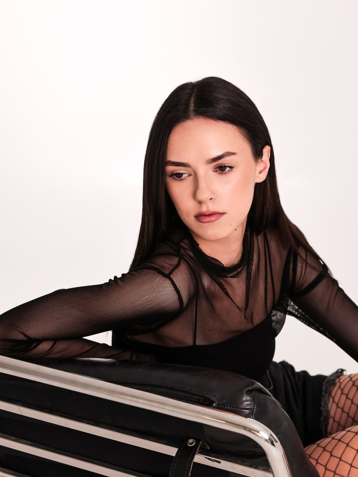 Leaning on a chrome chair in a sheer black top, against a white backdrop