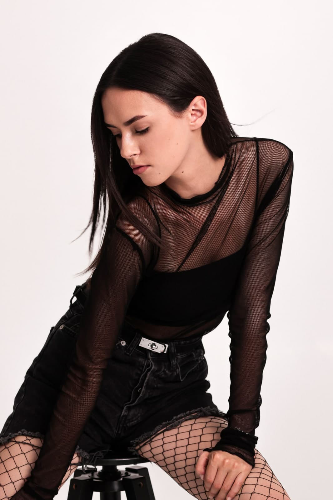 Sitting on a stool in a sheer black top with eyes lowered, white backdrop