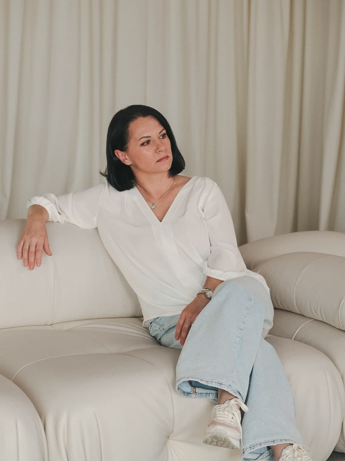 Leaning back on a white sofa in a white blouse and jeans, smiling softly