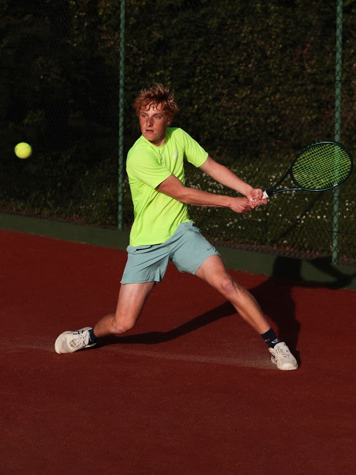 Two-handed backhand on a red tennis court in a neon-yellow T-shirt, the ball in the air