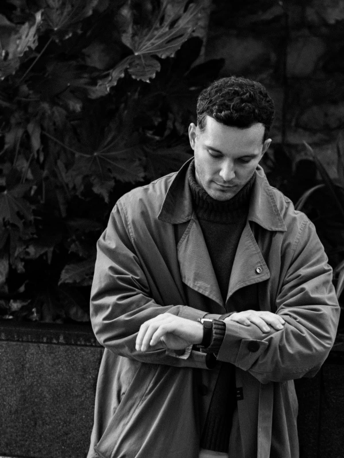 Black-and-white portrait checking a wristwatch, in a trench coat and roll-neck