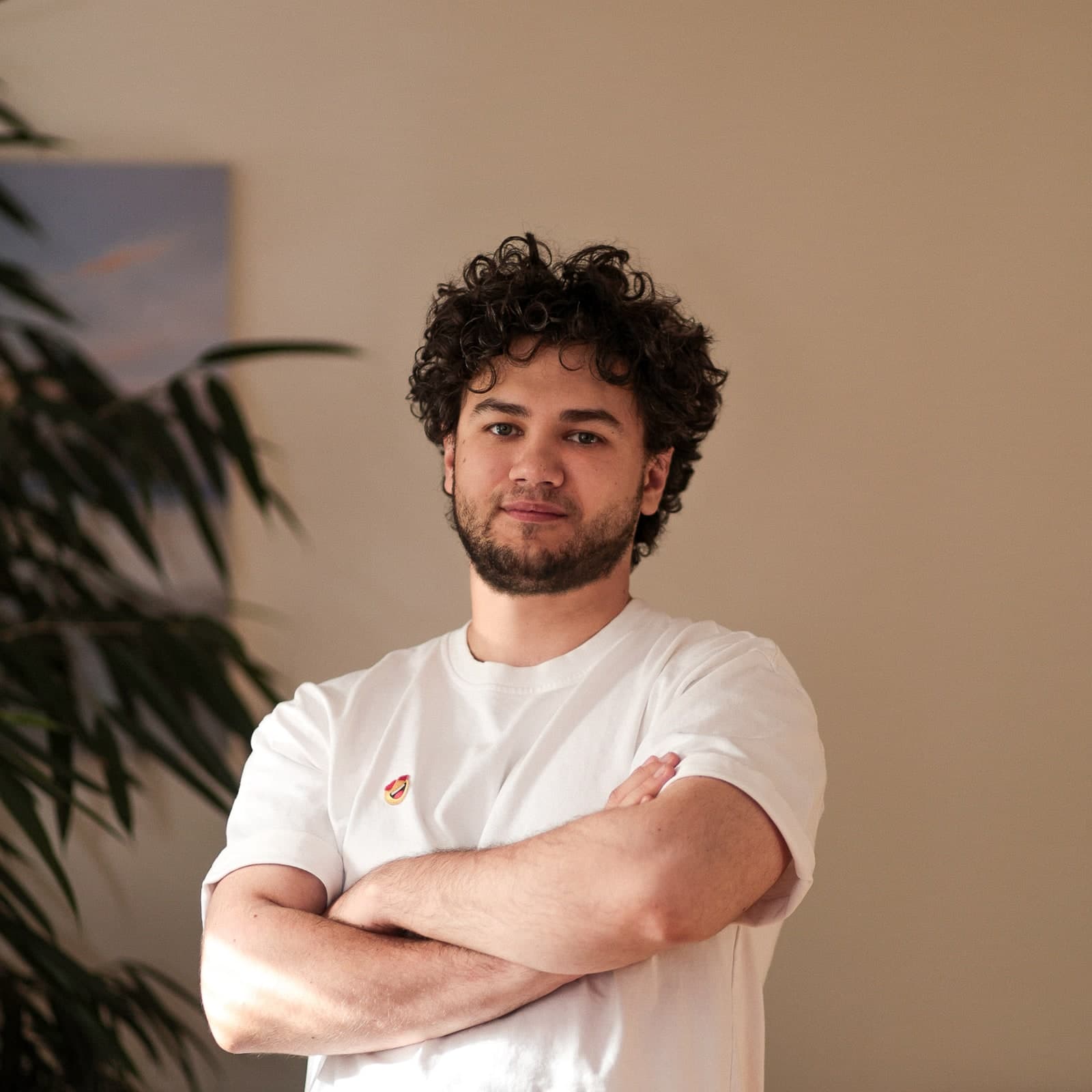 Relaxed portrait with curly hair and folded arms, in a white T-shirt beside a houseplant