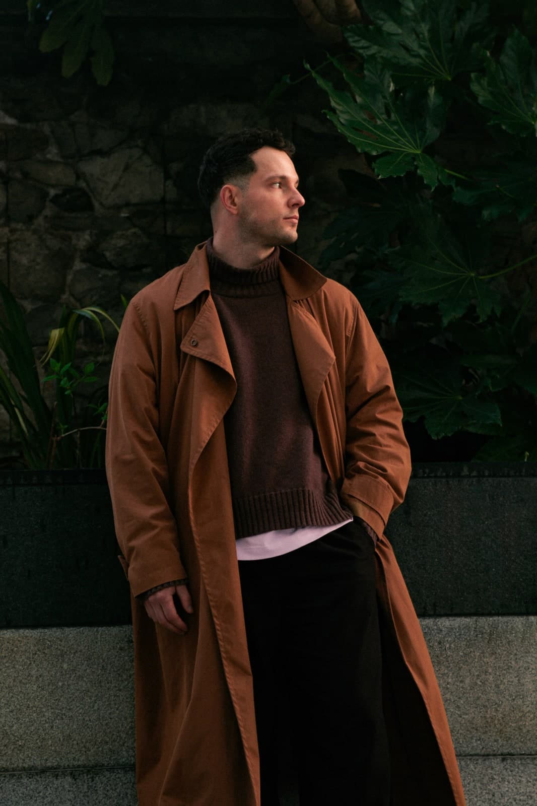 Leaning against a wall of tropical leaves in a long rust-coloured coat, looking away