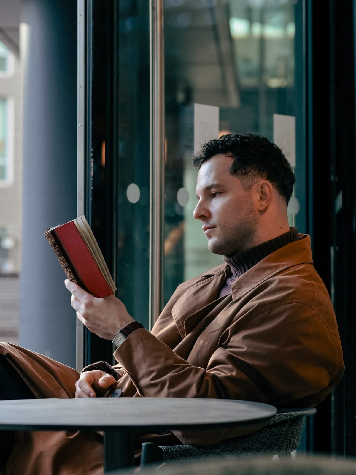 Reading a red book at a café table by a glass door, in a brown leather jacket