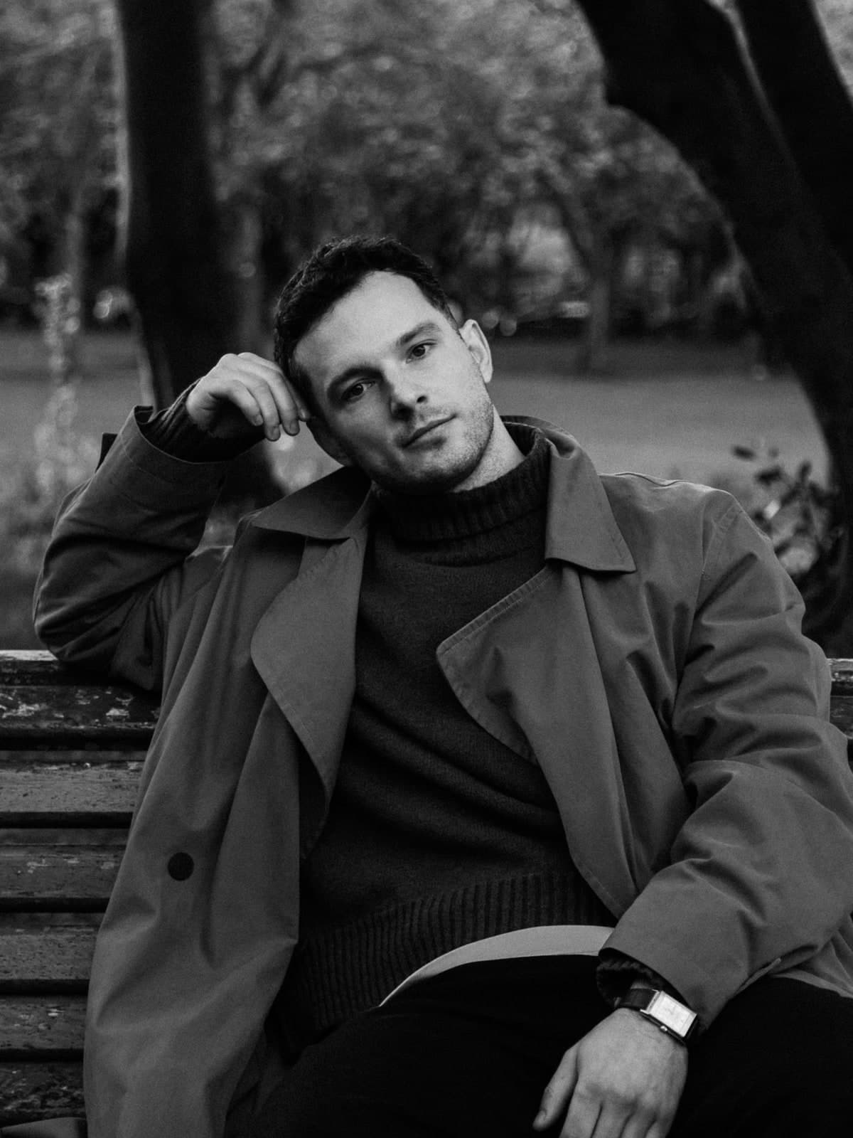 Black-and-white portrait on a park bench in a trench coat, head resting on one hand